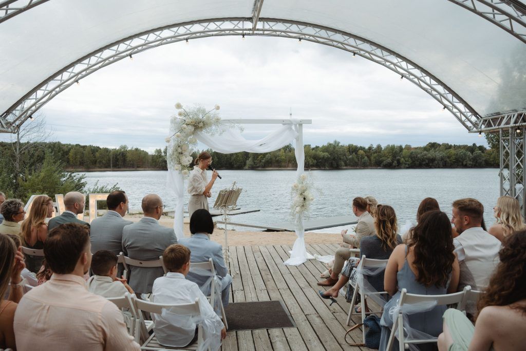 Hochzeitszeremonie in Neuss direkt am See mit Rednerin und Gästen Freie Trauung am Sandhofsee mit Blick auf das Wasser und elegantem Traubogen